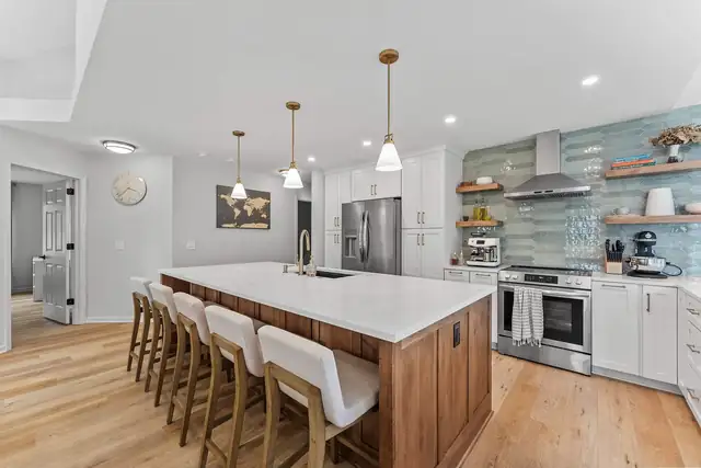 Kitchen by Keselman Construction Group with a long wood island, white quartz top, upholstered counter stools and a glass tile backsplash with open shelves